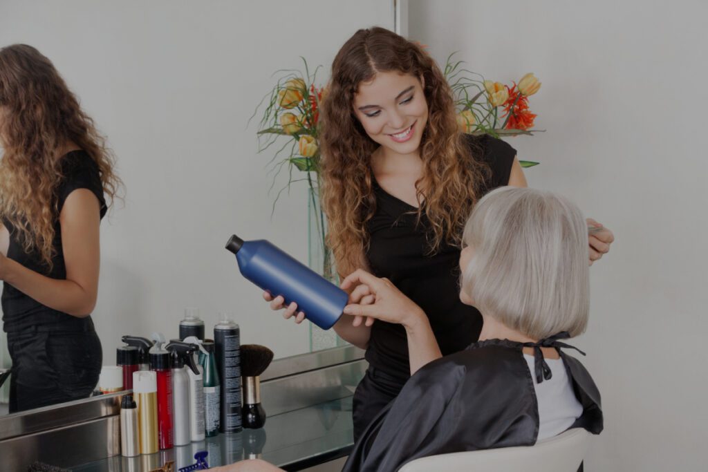 Eine lächelnde junge Coiffeuse mit lockigem Haar hält eine blaue Haarpflegeflasche und berät eine ältere Frau mit silbergrauem Haar, die auf einem Stuhl im Salon sitzt.
