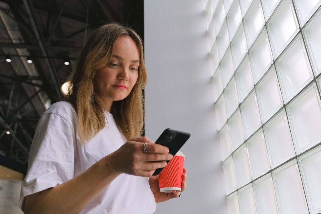 Eine Froschperspektive einer jungen Frau mit blonden Haaren in einem weissen T-Shirt, die auf ihr schwarzes Smartphone blickt, während sie einen roten Kaffeebecher zum Mitnehmen hält. Sie steht neben einer grossen Wand aus Glasbausteinen.